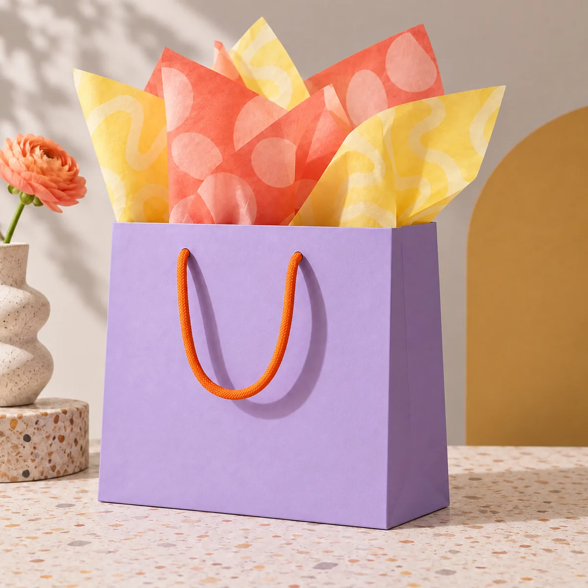 Fliederfarbene Tragetasche mit orangefarbenen Kordelgriffen und farbig gemustertem Seidenpapier, ohne weitere Verpackungsteile.